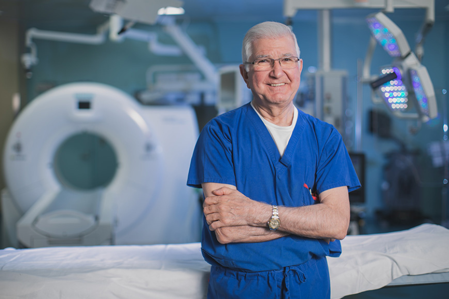 Dr. Tirone David poses in the surgical room in his blue surgical attire, smiling, with his arms crossed.