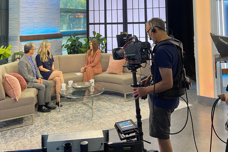 A behind the scenes look on a talk show with a cameraman filming a host talking to a UHN expert while siting on a couch and drinking coffee.